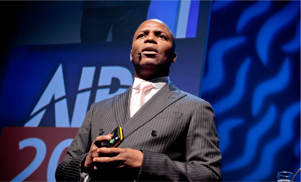 Thebe Ikalafeng speaking on stage in a pinstripe suit and headset microphone, lit against a blue backdrop